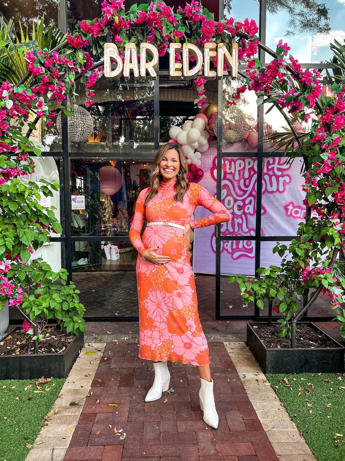 woman in floral dress holding baby belly in front of a sign that says bar eden