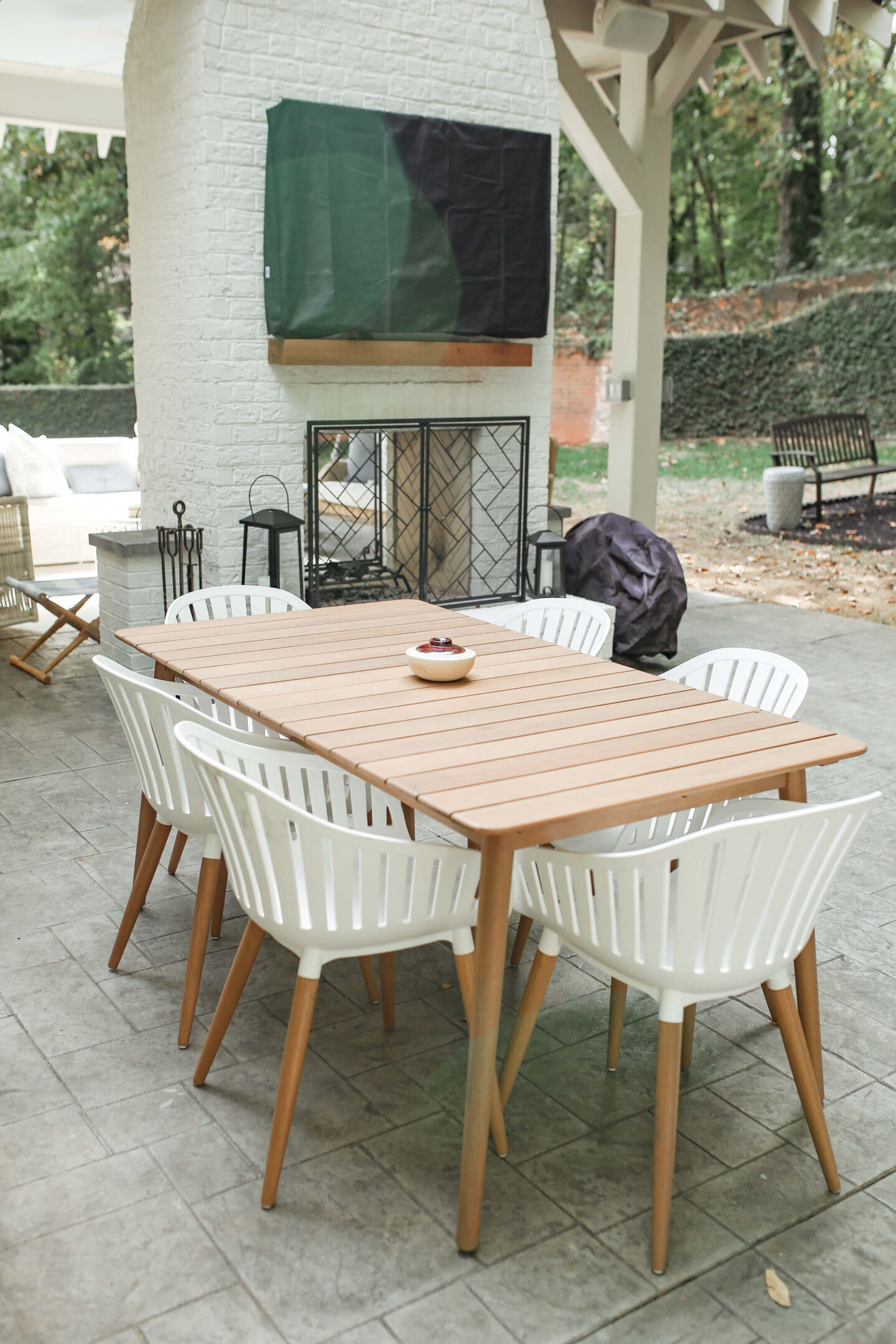 brown outdoor dining table with white dining chairs and outdoor fireplace in background