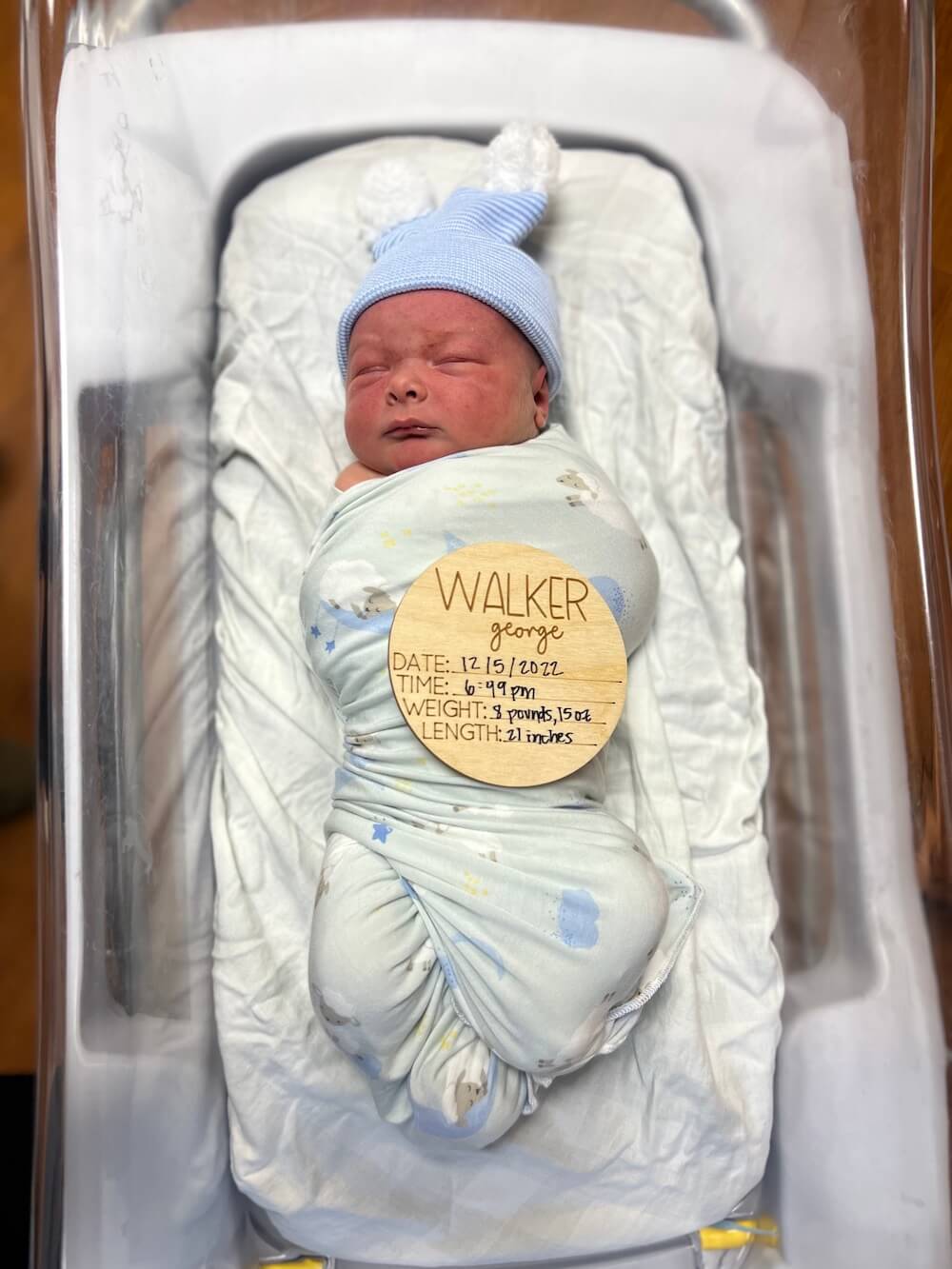 newborn baby swaddled in hospital bed with small round wooden sign that says Walker George