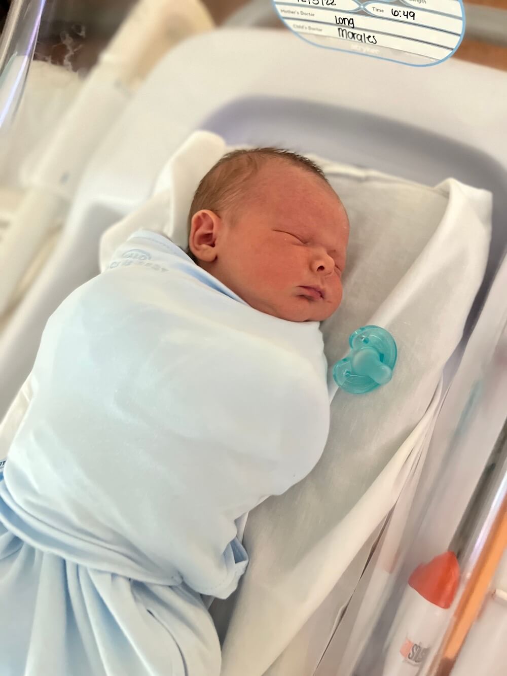 little boy swaddled in crib at hospital with pacifier laying next to him