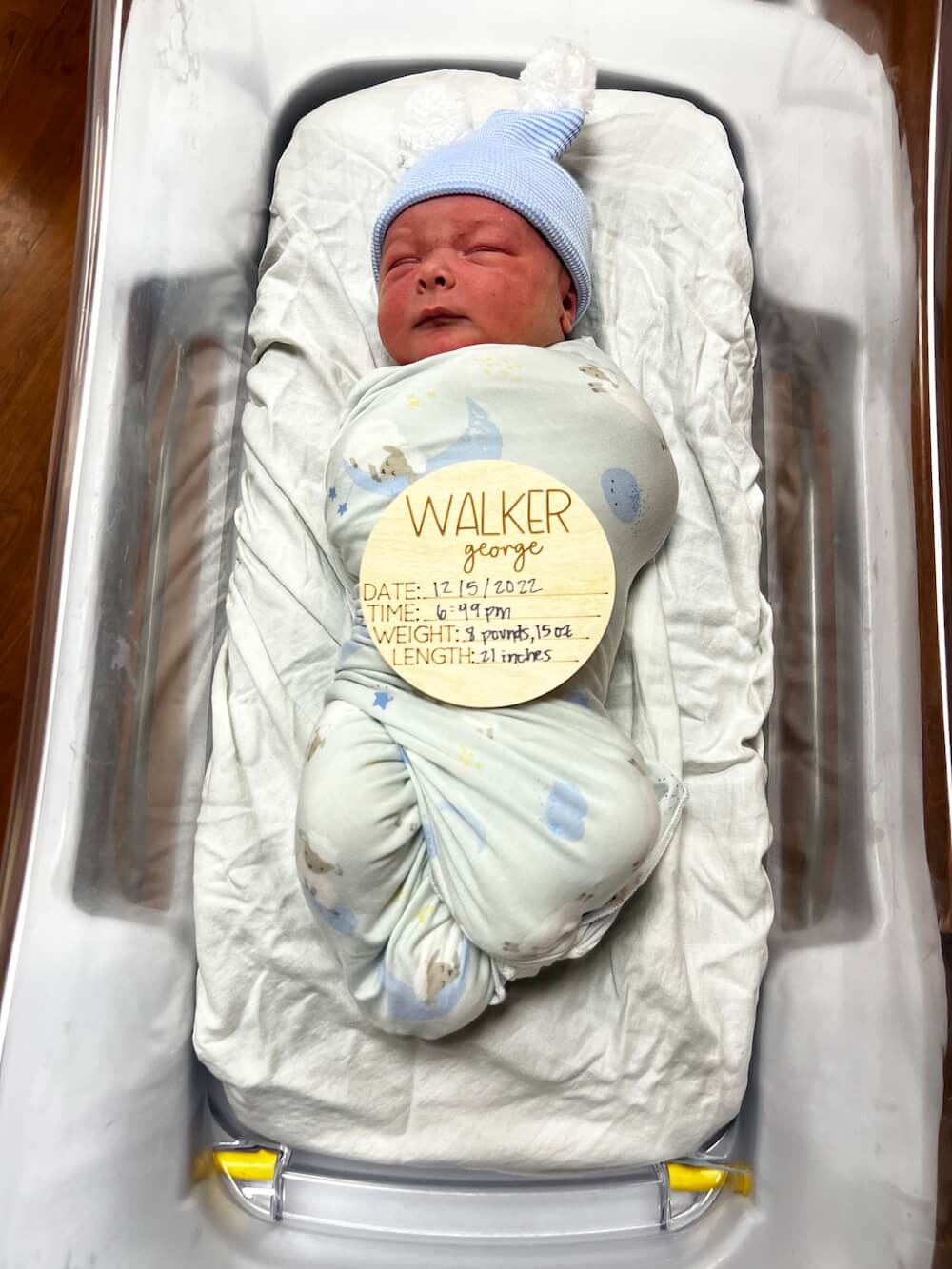 newborn baby swaddled in hospital bed with small round wooden sign that says Walker George