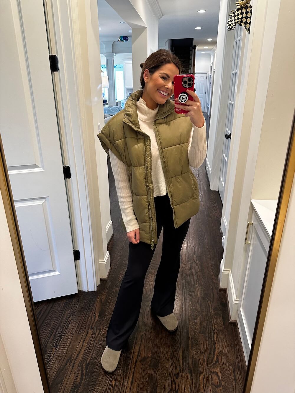 woman in tan puffer vest with jeans taking a mirror selfie